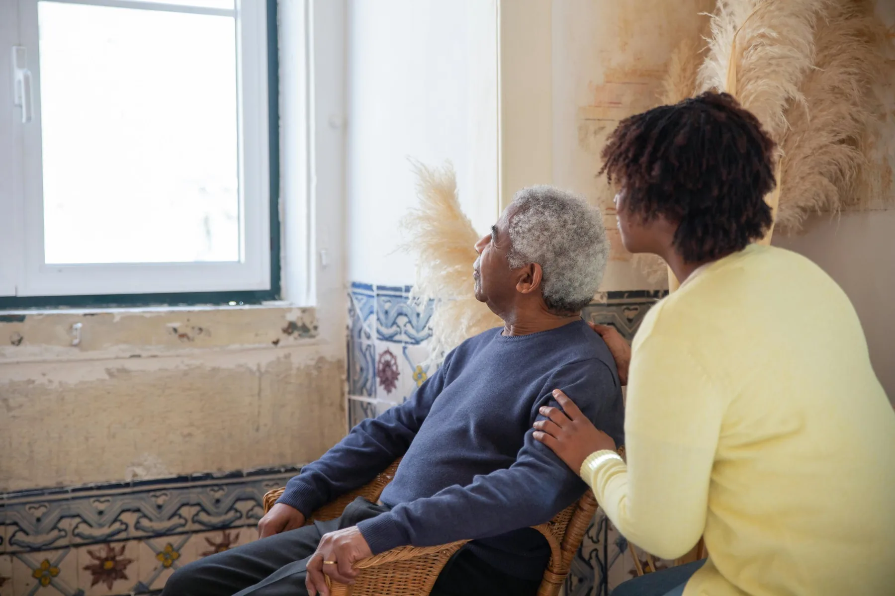 An elderly man seated at home, a younger woman beside him with her hand gently on his shoulder, both looking toward the window
